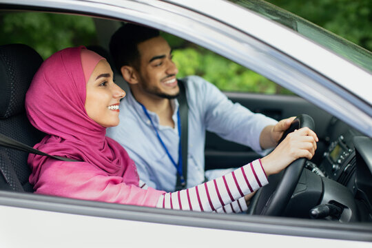 Caring Arab Husband Teaching His Muslim Wife In Hijab To Drive Car, Young Middle Eastern Man Helping Islamic Spouse In Driving Vehicle, Holding Steering Wheel And Smiling, Side View Shot