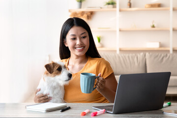 Cheerful young asian lady freelancer having coffee break while working online from home, sitting at...