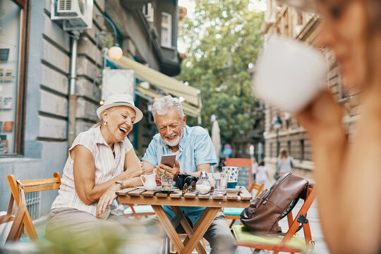 Senior couple laughing over smartphone at city sidewalk cafe