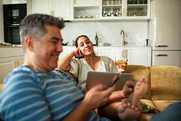 Couple relaxing on sofa with wine and devices at home kitchen