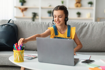 E-learning. Portrait of smiling little female student wearing headset using laptop computer in living room at home, taking pen from pencil holder, ready to take notes or draw. Homeschooling