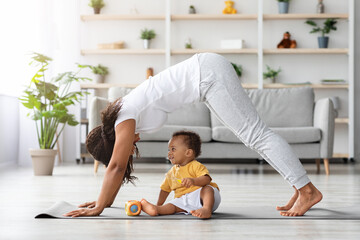 Young black mother practicing yoga at home with her infant son, standing in dog pose while training on fitness mat in living room, making back stretching exercise and smiling to her baby, copy space © Prostock-studio