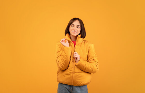 Cheerful young woman wearing and zipping up warm jacket, smiling and looking aside at free space, standing over orange studio background, copy space