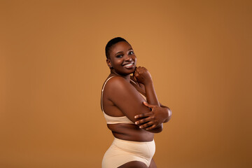 Plus Size Model. Profile Portrait Of Half-Turned Smiling African American Lady In Underwear Standing Over Brown Studio Background, Looking At Camera, Female In Lingerie Hugging Herself And Posing © Prostock-studio