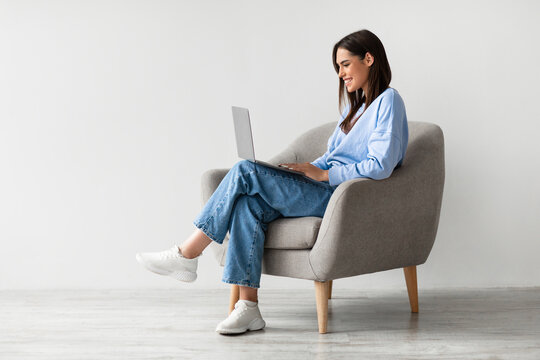 Smiling young woman in casual wear working online, sitting in armchair and using laptop against white studio wall, copy space. Cheerful Caucasian lady surfing internet on portable pc