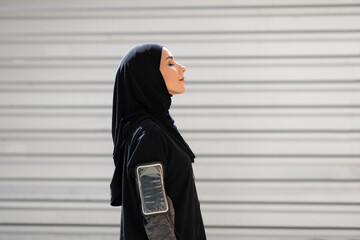 Tired young arab woman in hijab with phone on shoulder resting after exercise on gray wall background, profile. Outdoor sports, body care, training, ad and jogging. Health and body care due covid-19