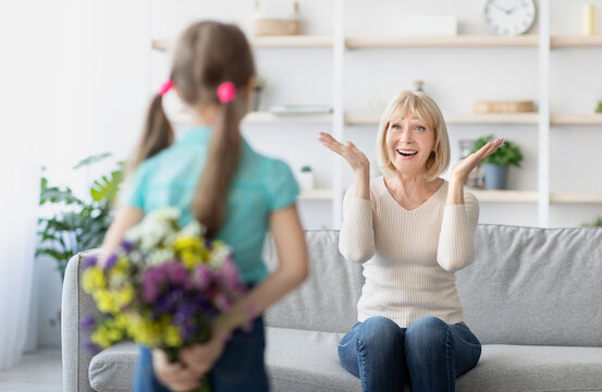 Happy Family. Rear view of little girl holding and hiding bouquet of fresh flowers behind her back, making surprise for exicted mature lady who sitting on couch in living room, selective focus