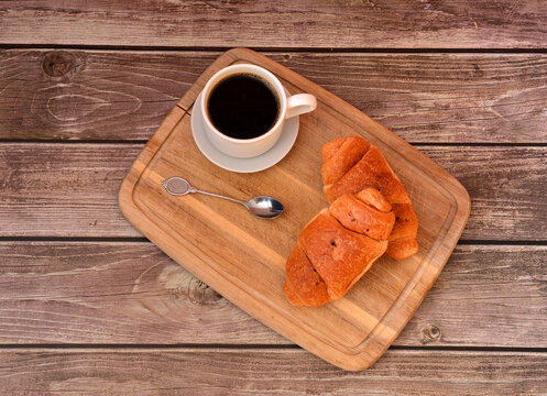 Wooden tray with two fresh croissants, a cup of hot black coffee and a tablespoon on a wooden table.