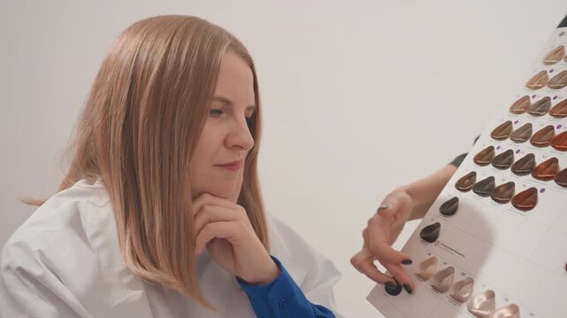 Caucasian woman choosing hair color swatches in modern salon, stylist presents sample board while client studies shades with thoughtful expression and gentle smile, closeup on hair texture