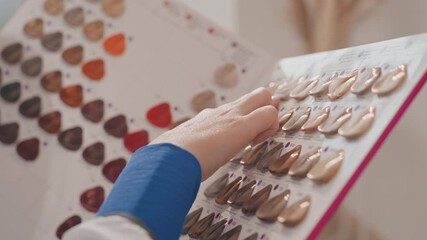Hands selecting nail polish swatches on board. Closeup of tattooed client hand and manicurist examining neutral and nude shades, glossy and matte finishes, organized palette on display, warm natural
