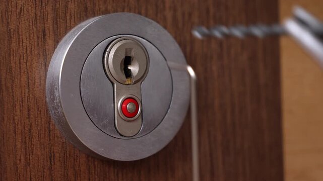 A closeup view shows lock picking tools being used on a modern round silver door lock set in a wooden surface, illustrating the process of manual lock picking