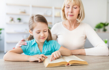 Naklejka premium Portrait of loving caring mature grandmother teaching cute little girl how to read, sitting at desk with interesting book, spending time together at home in living room. Preschool education concept