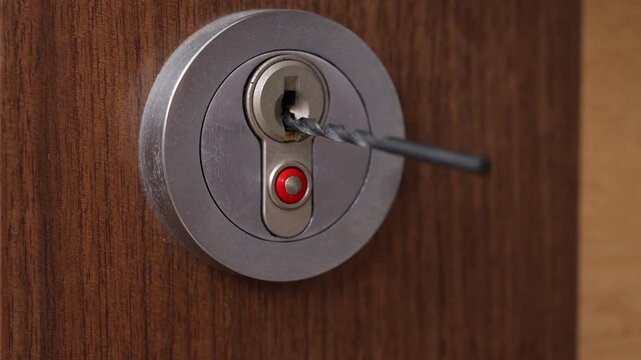 Close-up of a metal door lock with tools inserted into the keyhole, suggesting a lock picking attempt using a pick and tension wrench on a brown wooden door