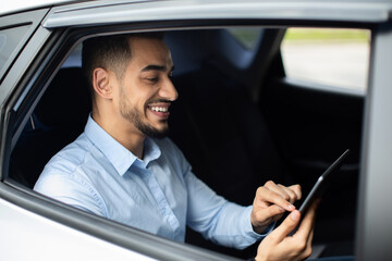 Cheerful middle eastern businessman checking emails while going to airport by taxi, sitting on car back seat and using digital tablet, closeup photo, shot from outside, side view, copy space