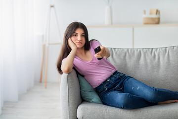 Indian teen girl with remote control watching TV on sofa at home, copy space. Female adolescent switching channels on television, feeling bored with movie or program on weekend
