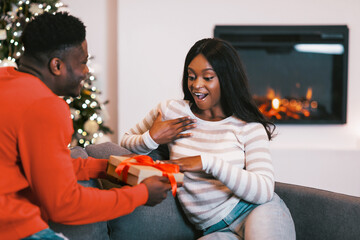 Obraz premium A man hands a wrapped gift to a woman sitting on a couch. She shows surprise and joy as they enjoy a moment together by a warm fireplace in a festive setting.