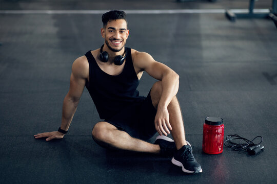 Portrait Of Handsome Arab Male Athlete Relaxing On Floor At Gym After Training, Young Muscular Middle Eastern Sportsman Smiling At Camera, Enjoying Bodybuilding And Healhy Lifestyle, Copy Space