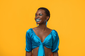 Obraz premium Attractive millennial african american woman with short hair in stylish blue outfit posing on yellow studio background, looking at copy space and smiling, wearing braces and bright makeup