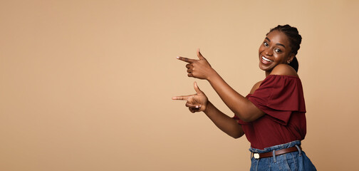 Cute millennial african american woman in stylish casual outfit pointing with both hands at copy space and smiling at camera, panorama, beige studio background. Advertisement, text concept © Prostock-studio