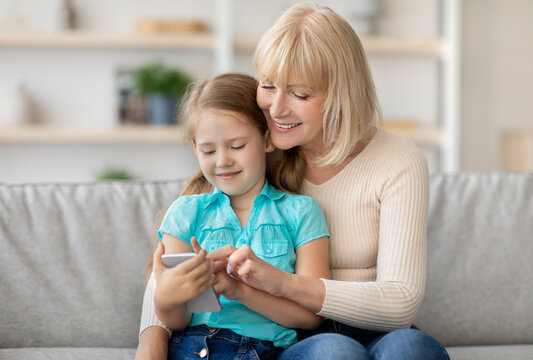 Portrait of smiling senior grandma and cute little kid granddaughter using cellphone watching cartoons together, happy family playing game, sitting on couch hugging and looking at screen