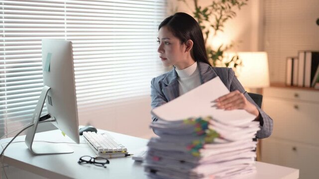 A woman is sitting at a desk with a stack of papers in front of her. She is looking at the papers and she is working