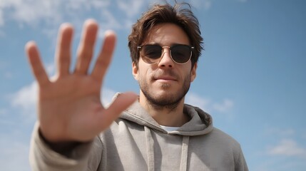 A man wearing sunglasses and a casual hoodie raises his open hand forward signaling a stop or greeting against a bright blue sky filled with