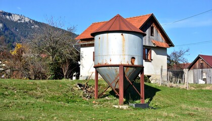 A rustic scene showing a metal storage container near a house with a red tiled roof. Green grass covers the foreground. Mountains are in the background