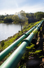 Two large green industrial pipes run parallel along a riverbank. This image represents infrastructure for water supply, sanitation, or oil and gas transportation, 