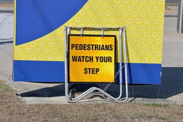 Pedestrians Watch Your Step sign mounted on a metal frame in front of a yellow and blue board