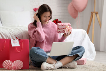 Beautiful young woman with laptop, gifts and shopping bags video chatting in bedroom. Valentine's...