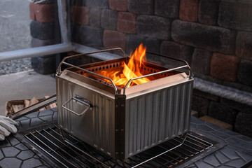 Stainless steel portable camping fire pit with burning wood fire, bright orange flames, cozy winter camping atmosphere, brick wall background.