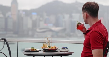 Man in red polo shirt drinking cocktail at rooftop bar facing Hong Kong skyline. Luxury travel, urban lifestyle, fine dining, vacation, city break, tourism.