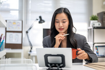 Asian businesswoman or office worker with a pen under chin holding coffee looking at tablet on table