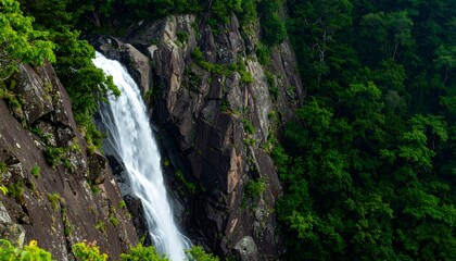 Fototapeta premium A scenic waterfall cascades down a rocky cliffside. Lush green foliage covers the right side. The waterfall appears white and forceful