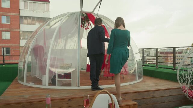 Rooftop bubble dome intimate date setup with couple entering clear igloo draped in red fabric, man hangs garland and lights while woman in green dress follows, cozy chairs and small table inside, city