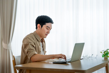 Happy young Asian man working on laptop in living room. Freelance worker male sitting at desk writing notes while watching webinar, studying online