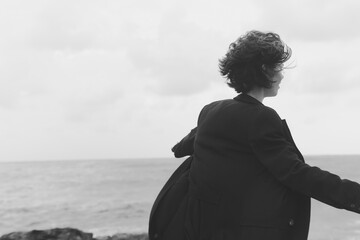 Figure in a dark coat stands at the wind swept coast, back to the camera, facing the sea and...