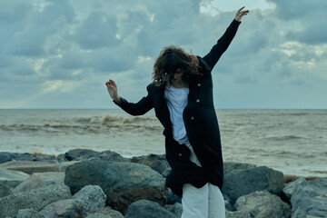 young woman coastal scene windy day rocky shore coat white outfit moves with wind joyful mood outdoor fashion style against dramatic ocean backdrop