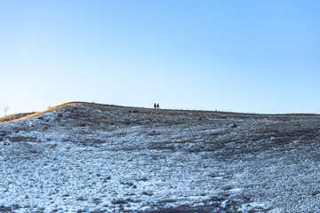 Two hikers stand on the snowy crest of a hill against a clear blue sky on a bright winter day. The low sun angle illuminates the golden dry grass on the upper part of the hill.