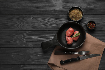 Frying pan with raw sausages, cutlery, bowls of dried herbs and peppercorns on black wooden...
