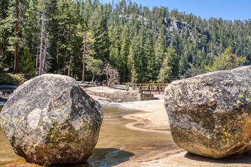 Two large rocks are on a beach near a body of water. The rocks are surrounded by trees and a...