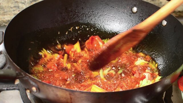 vibrant red saut&eacute; (sofrito) being stirred with a wooden spoon in a dark wok. The dish features tomatoes and peppers infused with annatto (onoto) oil, creating a rich base for traditional stews