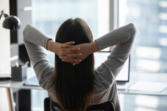 Office relax moment. Back view young woman corporate worker student take break for serene rest by laptop sit on chair in comfortable pose with fingers clasped behind head unwind distracted from screen