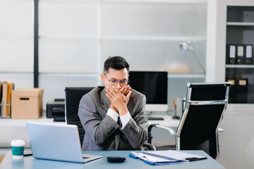Tired businessman sitting at office desk with laptop, feeling stressed and overworked under tight deadlines