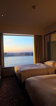 Modern luxury hotel room with twin beds. Large window view of ocean and city skyline. Warm golden hour sunlight. High-end travel and hospitality concept Vertical.