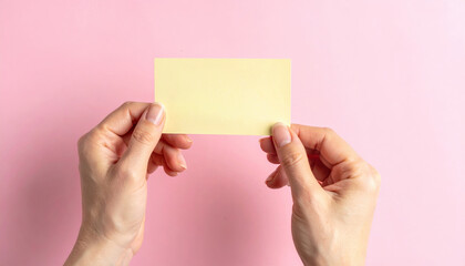 Two hands holding a blank yellow rectangular sticky note against a pink wall