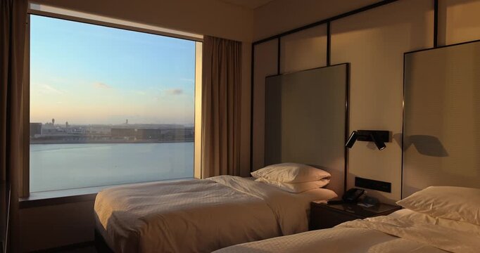 Modern hotel room with twin beds. Large window overlooks water and city at sunset. Warm golden hour lighting. Luxury hospitality and travel concept.