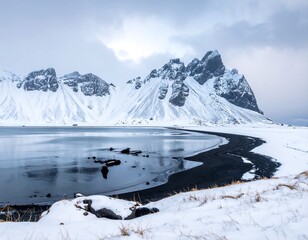 A dramatic winter scene of a black sand beach, icy lake, and snow-capped mountain range under a cloudy sky