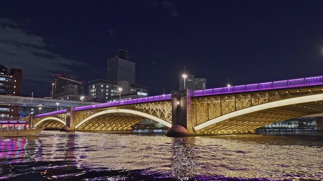 A wide nighttime view of the golden-lit arches of Kuramae Bridge with a purple-lit upper rail, as an illuminated tour boat glides across the Sumida River.