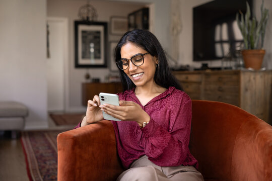 Positive young Indian woman in glasses typing on smartphone at home, relaxing in armchair, enjoying online communication, fast Internet connection, using smart technology on mobile phone